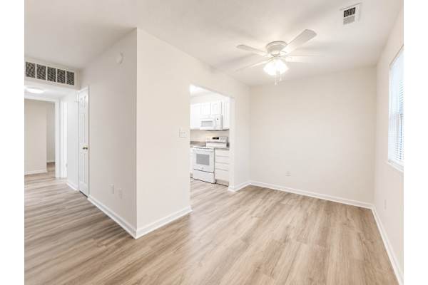 A room with a ceiling fan and light, wooden floors, and a kitchen area in the background.